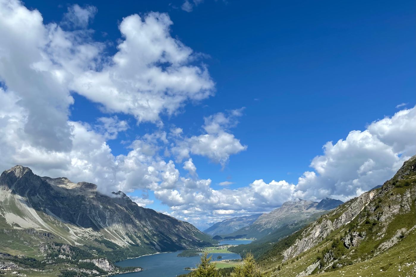 A wide mountain valley in summer: a long lake far below, green slopes and grey peaks, a blue sky full of white clouds